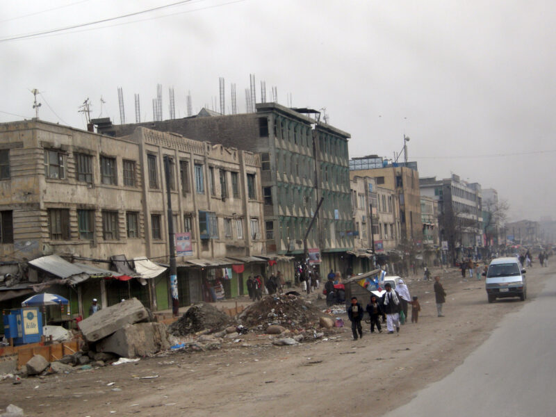 A dreary streetside scene in downtown Kabul. Concrete buildings and various detritus line a dusty road.
