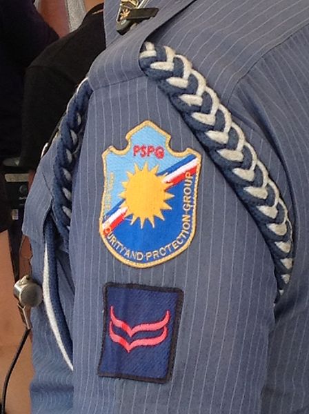 The shoulder of a Philippine National Police uniform. There is a braid around the shoulder, a badge for the Security and Protection Force, and a blue square badge with two red chevrons.