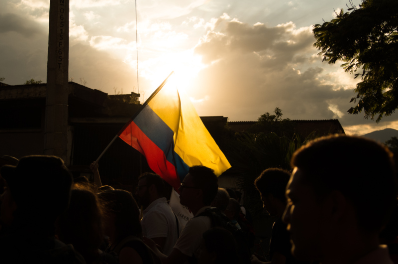 A crowd of people, silhouetted and backlit by a setting sun, hold aloft a Colombian flag.