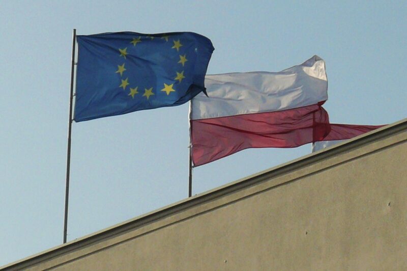 The EU and Polish flags, photographed atop a building against a clear sky.