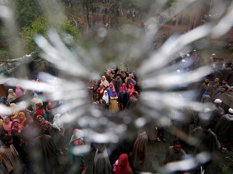 A photograph of a protest against Indian occupation in Kashmir. The photo is taken behind glass, with the people in the street below framed by a hole shattered through it, as if it had been pierced by a bullet.