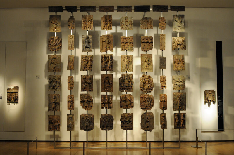 A wall of Benin bronzes displayed at the British Museum. These bronze plates depict various figures and are hung on poles in a neat array.