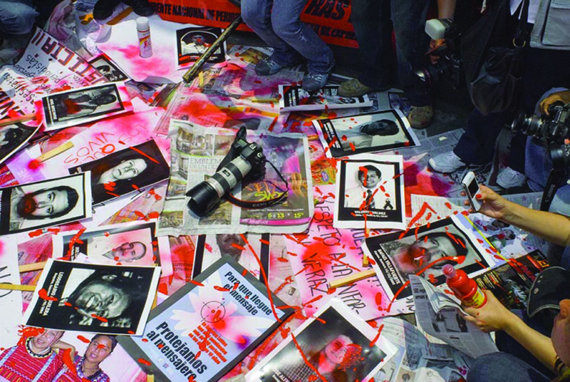 A photograph from a Mexican protest over violence against journalists in 2010. Newspapers, signs, and posters (presumably of deceased journalists) lie in a pile, spraypainted red to evoke blood spatters. A camera has been left on top.