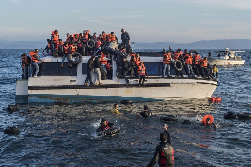 Syrian and Iraqi refugees arrive from Turkey to Skala Sykamias, Lesbos island, Greece in 2015. Many, many people, most in life jackets, crowd on top of every standable surface on a small boat. More appear to be packed tightly inside, and a few others are scattered in the water.