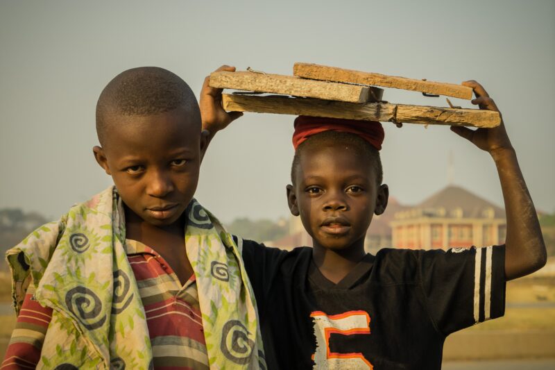 Two Nigerian boys, photographed against a gray sky. One wears a striped polo with green sun- and flower-printed fabric draped over his shoulders. The other wears a sports jersey and a red hat and carries wood over his head.