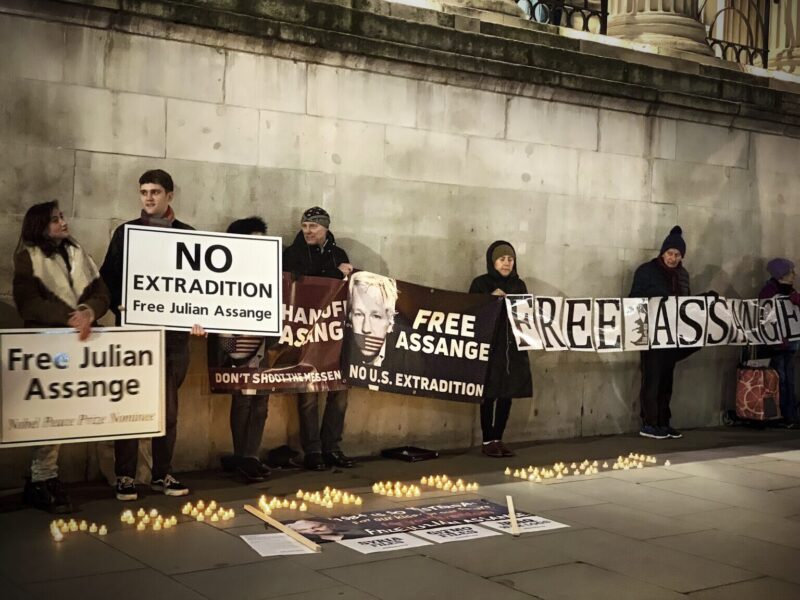 People gather against a wall during a vigil for Julian Assange at Trafalgar Square. The people are holding signs with the slogans "Free Assange" and "No extradition."