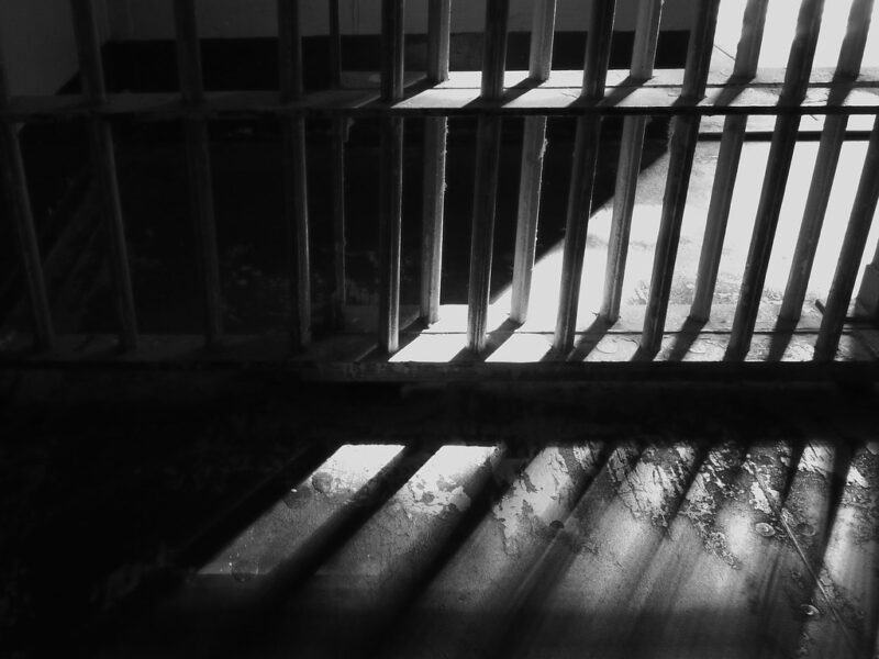 A black and white photograph of a jail cell, taken from a low angle to show only the bottom of a jail door. Light streams through the bars in stripes.