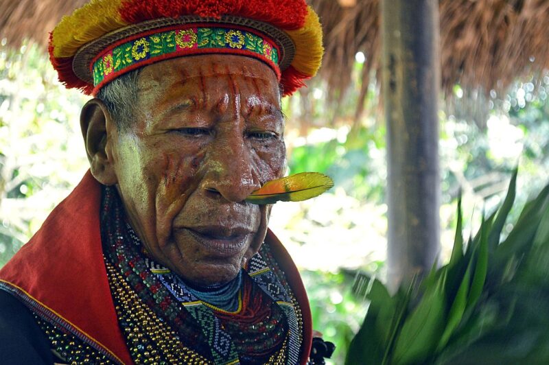 An elderly Cofán man, photographed in what appears to be a hut. He wears many colorful beads around his neck and an intricately embroidered headdress with a flat fringe radiating from around the forehead. There is a rainbow feather pierced through his septum.