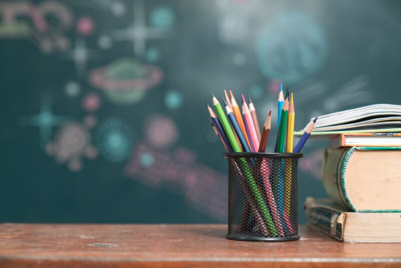A photograph of a school desk. A can of colored pencils sits next to a stack of books. A doodled-upon chalkboard is out of focus in the background.