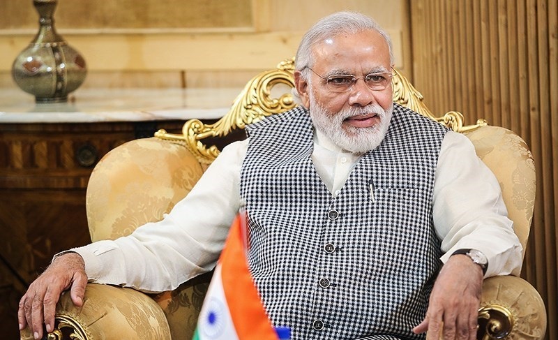 Prime Minister Narendra Modi, photographed sitting wide in a large armchair. What is visible of the room is beige and finely decorated. A small Indian flag is just visible in front of him.