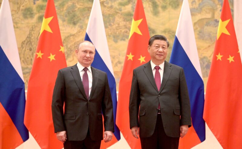 Vladimir Putin and Xi Jinping, photographed side by side in front of a row of alternating Russian and Chinese flags.