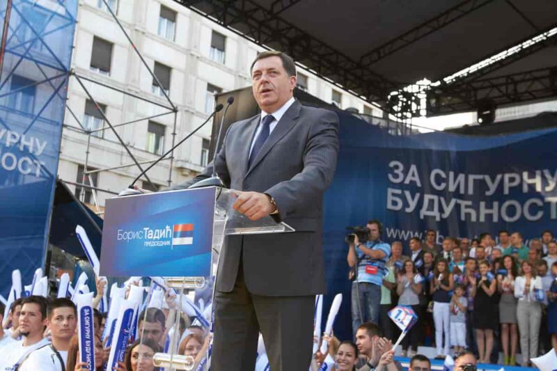 Milorad Dodik gives a speech at a Russian rally. He stands in front of Cyrillic signs and his lectern has a Russian flag on it.