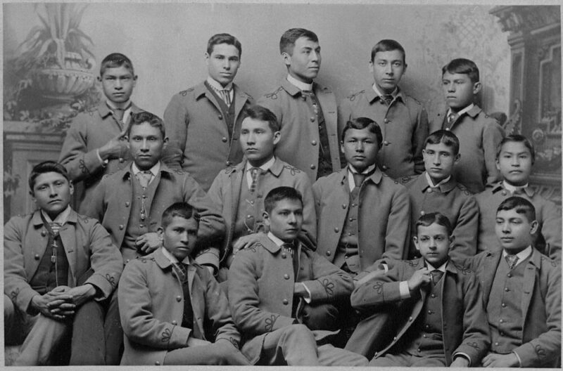 Students of the Carlisle Indian Industrial School, circa 1986. The students wear suits and identical, cropped haircuts. Their expressions are solemn and pained. The photo is in black and white.