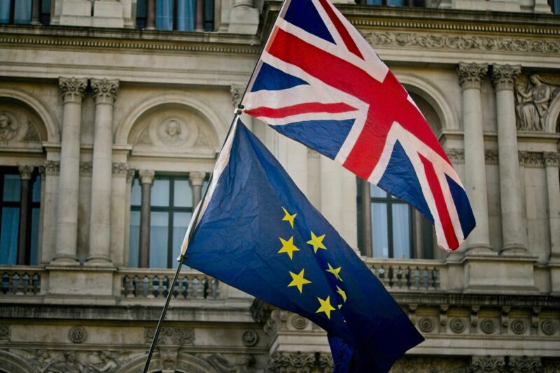 A photograph taken in London in 2018. A U.K. flag flies above an E.U. flag in front of a government building edifice.