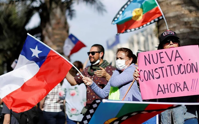 Protestors demonstrate, waving Chilean and Mapuche flags. A pink sign reads "NUEVA CONSTITUCIO´N AHORA" ("NEW CONSTITUTION NOW").