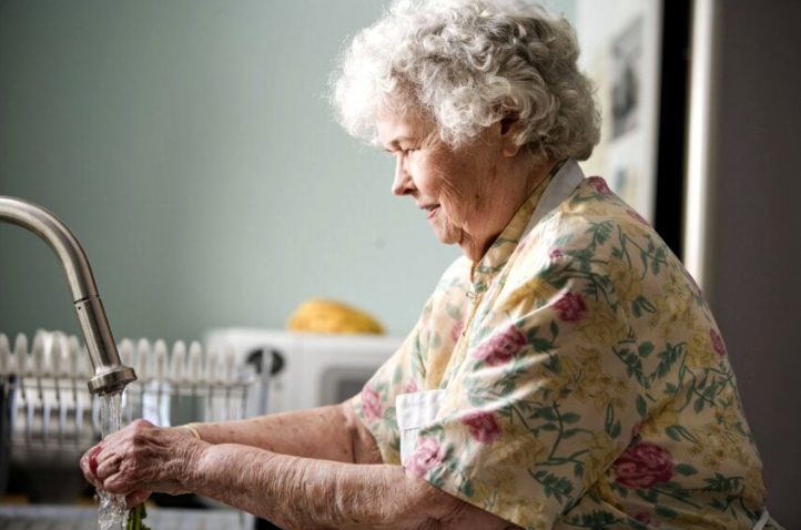 An old woman, seen from the side, standing at a sink. She wears a pale, floral shirt, and is washed out, photographed in weak light with a strong vignette and a pale background she fades into.