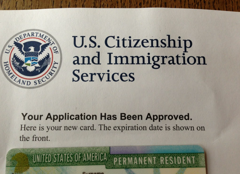 A document from U.S. Citizenship and Immigration Services notifies the recipient, "Your Application Has Been Approved." The top of a "green card" is visible at the bottom of the photograph.