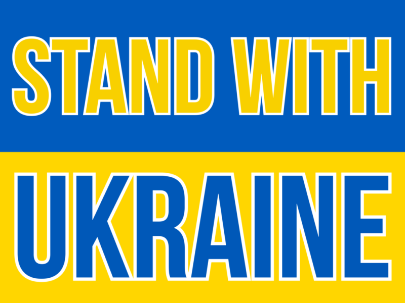 A poster of the Ukrainian flag. The words "STAND WITH" are written on the blue stripe in yellow, with "UKRAINE" concluding the sentence in blue over the yellow half.