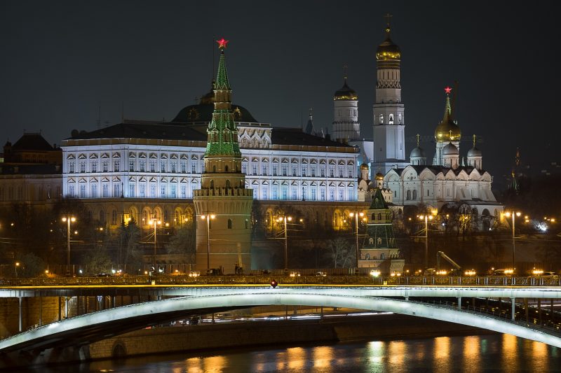 The Kremlin, photographed at night from the other side of a bridge. A red star tops the closest tower. The famous domed towers are in the top right, effectively creating a solid wall of towering building overlooking the river.