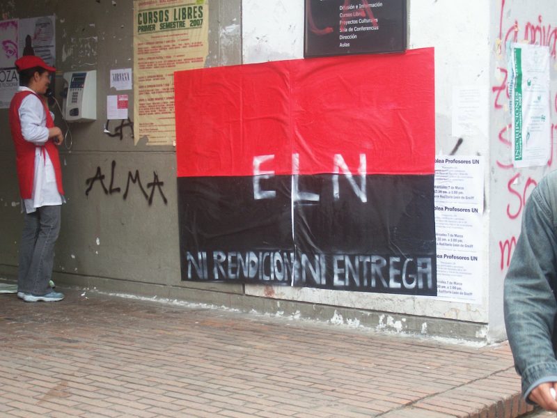 An E.L.N. guerrilla poster on a graffiti- and poster-covered wall. The poster is the E.L.N. flag, red and black with the letters ELN in white across the middle, with "Ni rendición ni entrega" – "Neither capitulation nor surrender" – written across the bottom. The word "ALMA" (heart; soul) is scrawled next to it in a different hand, part of another graffiti tag evidently covered by one of the other posters.