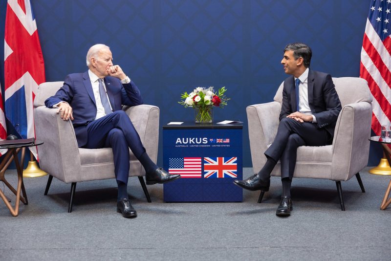 President Joe Biden and Prime Minister Rishi Sunak talk, photographed in armchairs bracketing a pedestal labeled AUKUS with all three nations' flags. The leaders sit opposite their flags, with Biden in front of the Union Jack and Sunak in front of the Stars and Stripes.