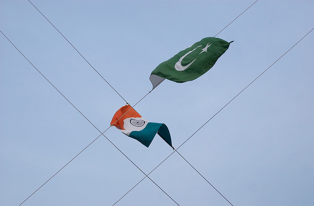 An Indian and Pakistani flag, photographed from below against a blue-gray sky. They're strung up on opposing, perpendicular lines crossing the camera field like barbed wire.
