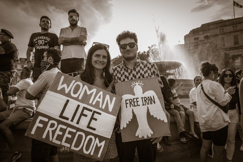 Protestors photographed in Trafalgar Square. A woman holds a striped sign reading "WOMEN LIFE FREEDOM". A man holds a sign with a dove and the words "FREE IRAN".