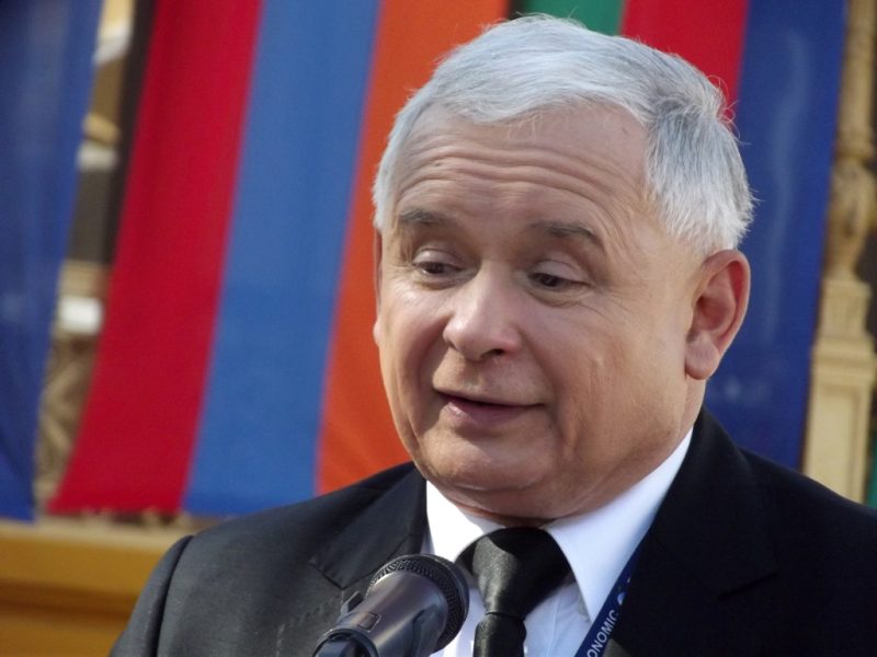 PiS Chairman Jarosław Kaczyński, photographed behind a microphone and in front of vertically red and blue-striped flags.